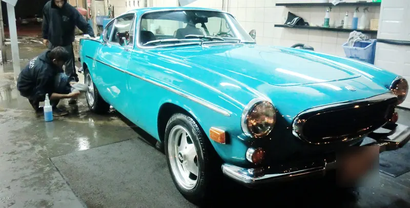 Two workers hand-washing a classic turquoise Volvo coupe on the wet floor of the wash bay